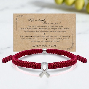 May include: A burgundy macrame bracelet with a silver ribbon charm. The bracelet is next to a card with the text "Life is tough, but so are you!" and the words "LOVE - HOPE - LIFE".