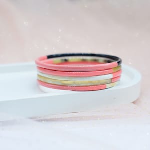 May include: A stack of five colourful bangles, featuring a pink, black, white, and gold colour scheme. The bangles are arranged on a white surface.