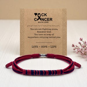 May include: A red and navy blue braided bracelet with a silver clasp. The bracelet is displayed with a card that reads "F*ck Cancer" in a heart shape, with the words "morse code" and a message of support.