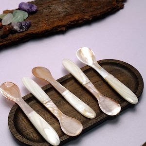 Puede incluir: Cinco cucharas de nácar dispuestas sobre una bandeja de madera oscura. Las cucharas tienen un brillo nacarado con un tono marrón claro en el cuenco y mangos blancos. La bandeja es de forma ovalada. El fondo es de color lila claro.