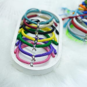 Peut inclure: Collection de bracelets tress&eacute;s color&eacute;s avec un charm en forme de ruban argent&eacute;. Les bracelets sont pr&eacute;sent&eacute;s dans un plateau ovale blanc sur une surface blanche et duveteuse. Les bracelets sont disponibles en plusieurs couleurs, dont rose, violet, vert, jaune, noir et bleu.
