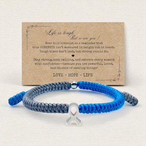 May include: A blue and gray braided bracelet with a silver ribbon charm. The bracelet is displayed in front of a card with the text "Life is tough, but so are you!" and the words "LOVE - HOPE - LIFE".