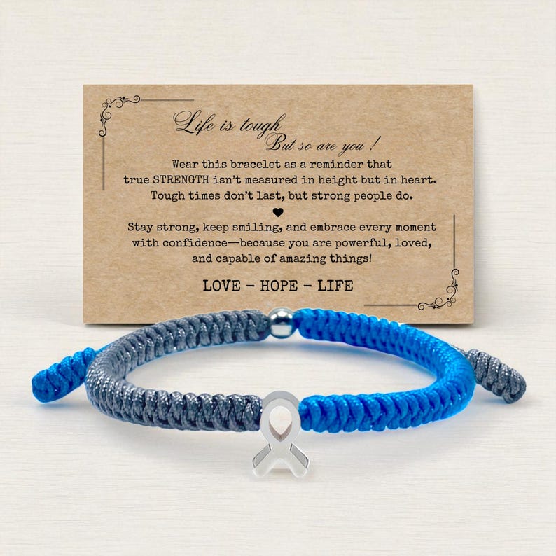 May include: A blue and gray braided bracelet with a silver ribbon charm. The bracelet is displayed in front of a card with the text "Life is tough, but so are you!" and the words "LOVE - HOPE - LIFE".