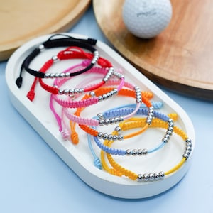 May include: A selection of colourful friendship bracelets with adjustable knots and silver-coloured beads. The bracelets are in various colours, including black, red, pink, orange, blue, and yellow, displayed on a white tray.