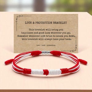 Peut inclure: Un bracelet rouge, blanc et argent avec une carte indiquant "LUCK & PROTECTION BRACELET". Le bracelet est fait de fils rouges et blancs tissés, avec des perles argentées au centre. La carte comprend un message sur le bonheur et la chance.
