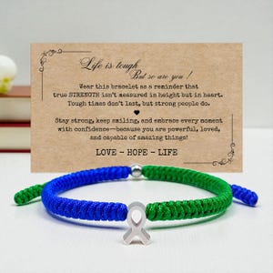 May include: A blue and green braided bracelet with a silver ribbon charm. The bracelet is displayed with a card that reads "Life is tough, but so are you!" and includes the words "LOVE - HOPE - LIFE".
