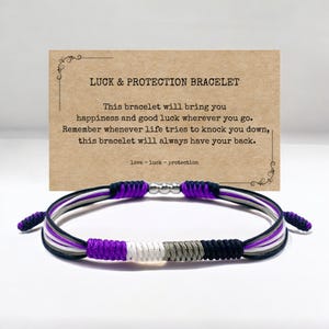 Op de afbeelding: Een gevlochten armband in paars, zwart, wit en grijs met zilveren kralen. De armband wordt geleverd met een kaart waarop "LUCK & PROTECTION BRACELET" staat en de tekst "Deze armband brengt je geluk en geluk waar je ook gaat. Denk eraan, wanneer het leven je probeert neer te halen, deze armband zal altijd voor je klaarstaan. liefde - geluk - bescherming"
