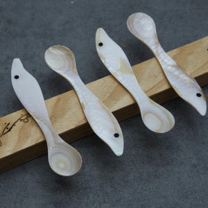 Puede incluir: Cuatro cucharas decorativas con forma de pez. Las cucharas están hechas de un material nacarado con un patrón de escamas de pez. Cada cuchara tiene un pequeño punto negro para un ojo. Las cucharas descansan sobre una base de madera.