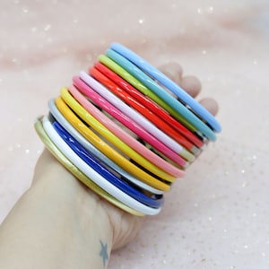 Könnte beinhalten: Ein Stapel bunter Armreifen in verschiedenen Blautönen, Grün, Rot, Rosa, Gelb und Weiß. Die Armreifen sind aus einem glänzenden, glatten Material gefertigt und übereinander gestapelt.
