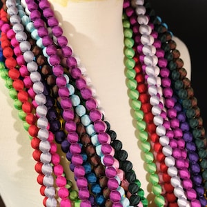 Może przedstawiać: Kolekcja kolorowych naszyjników z koralików w różnych odcieniach, w tym czerwonym, zielonym, fioletowym i niebieskim. Koraliki są okrągłe i wydają się być wykonane z tkaniny. Naszyjniki są ułożone na manekinie, prezentując różne opcje kolorystyczne.