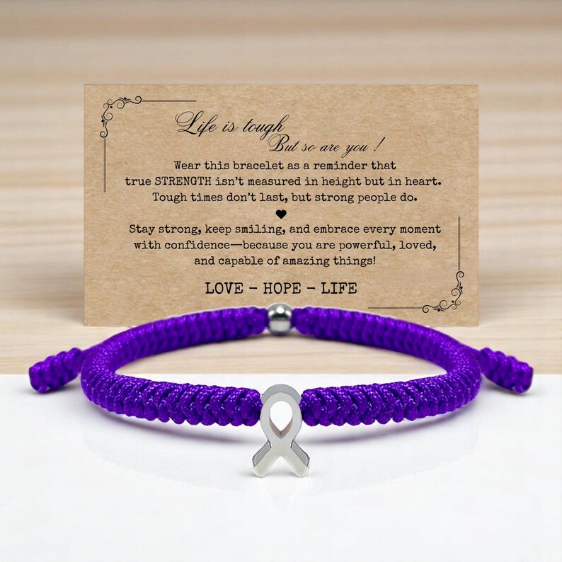Peut inclure: Un bracelet tress&eacute; violet avec un charm en forme de ruban argent&eacute;. Le bracelet est pr&eacute;sent&eacute; devant une carte avec le texte "Life is tough, but so are you !" et les mots "LOVE - HOPE - LIFE".