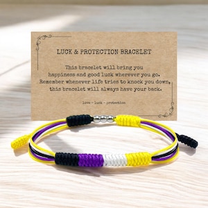 Könnte beinhalten: Ein mehrfarbiges Armband aus Schnur mit gelben, weißen, schwarzen und violetten Fäden. Das Armband hat silberne Perlen und eine Karte mit der Aufschrift "GLÜCK & SCHUTZ-ARMBAND. Dieses Armband bringt dir Glück und Zufriedenheit, wohin du auch gehst. Denk daran, wann immer das Leben versucht, dich zu Fall zu bringen, dieses Armband wird immer für dich da sein. Liebe - Glück - Schutz"