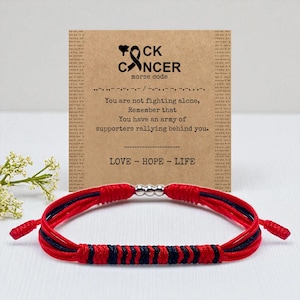 May include: A red and navy blue bracelet with a silver bead clasp. The bracelet is displayed in front of a card with the text "FOCK CANCER" and a message of support. The card also includes the Morse code for the word "cancer".