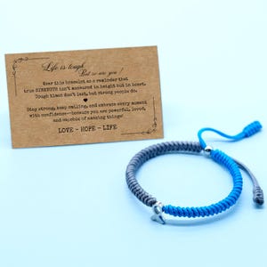 May include: A gray and blue braided bracelet with a silver charm in the shape of a ribbon. A card with text that reads "Life is tough but so are you!" is in the background. The card also includes the words "LOVE - HOPE - LIFE".