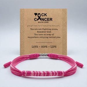 May include: A pink and white braided bracelet with a silver bead clasp. A card behind the bracelet reads "F*ck Cancer" with a Morse code message and the words "LOVE - HOPE - LIFE".