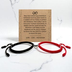 Könnte beinhalten: Passende Freundschaftsarmbänder, eines in Schwarz und eines in Rot, mit einem herzförmigen Magnetverschluss. Die Armbänder werden mit einer Karte mit dem Text "Keep this matching protection bracelet close to your heart as a symbol of our bond..." und den Worten "LOVE - LUCK - PROTECTION" präsentiert.