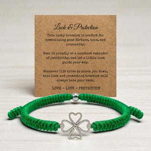 May include: A green braided bracelet with a silver four-leaf clover charm. The bracelet is displayed with a card that reads "Luck & Protection". The card's text describes the bracelet as a symbol of good fortune, hope, and prosperity.