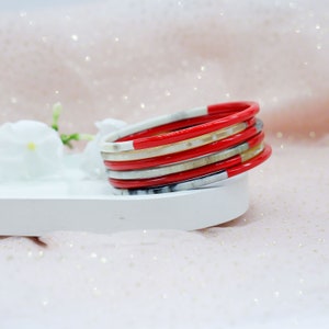 May include: Stack of colorful bangles. The bangles are red, white, and silver. The bangles are stacked on a white tray with a white flower in the background. The background is a soft pink color.