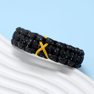 Peut inclure: Bracelet en paracorde noir avec un accent en forme de X jaune.
