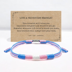 May include: A blue, pink and white braided string bracelet with silver beads. The bracelet is on a brown card with the text "LUCK & PROTECTION BRACELET" and a message about the bracelet bringing happiness and good luck.