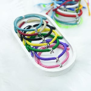 May include: A selection of brightly coloured braided cord bracelets with silver-tone details, presented in a white oval tray. The bracelets are in various colours, including pink, purple, green, yellow, black, and red. More bracelets are visible in the background.