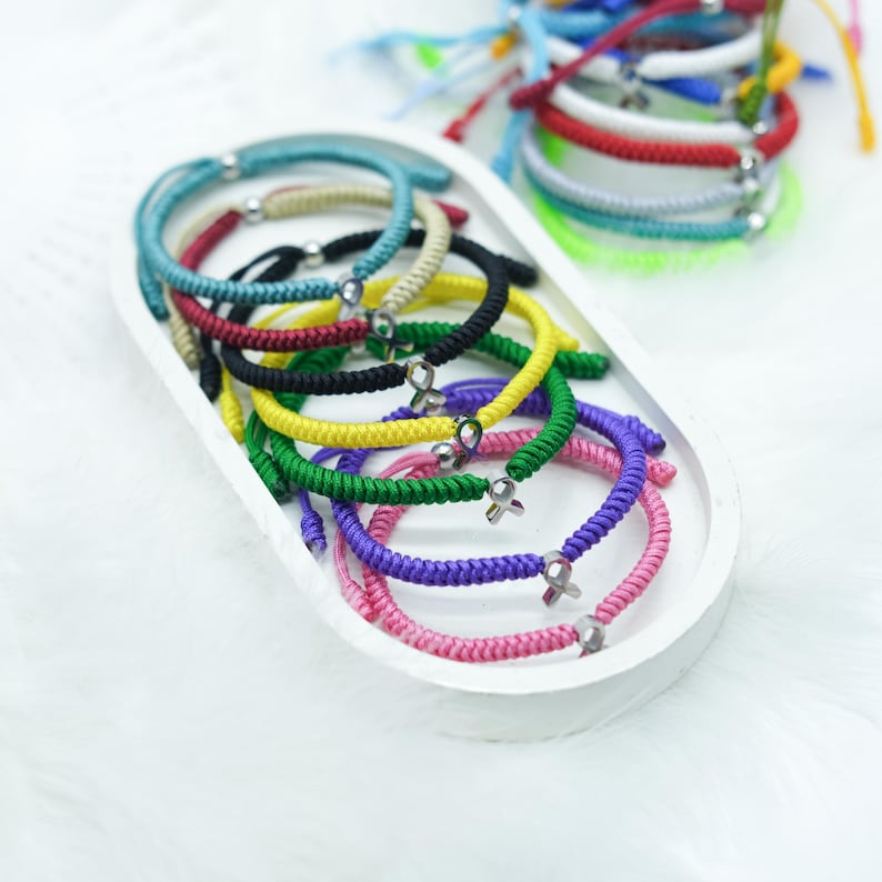 Peut inclure: Une collection de bracelets en cordon tress&eacute; color&eacute; avec des accents argent&eacute;s, pr&eacute;sent&eacute;e dans un plateau ovale blanc. Les bracelets sont de diff&eacute;rentes couleurs, dont rose, violet, vert, jaune, noir et rouge. D'autres bracelets sont visibles en arri&egrave;re-plan.