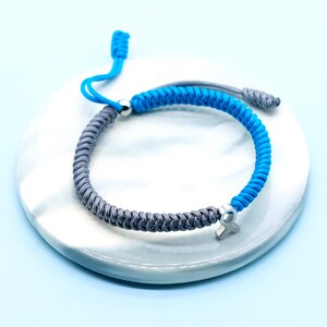 May include: A blue and gray braided bracelet with a silver cross charm. The adjustable bracelet is made of a woven cord and features a sliding knot closure. The bracelet is displayed on a white, marbled surface against a light blue background.