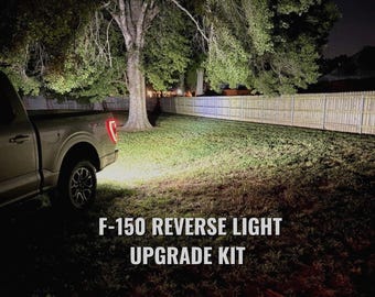Puede incluir: Un camión F-150 plateado con las luces de marcha atrás encendidas, iluminando la hierba. El texto "F-150 REVERSE LIGHT UPGRADE KIT" se muestra en blanco en la parte inferior de la imagen. La escena está ambientada de noche.