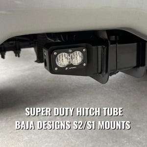 May include: A black metal hitch tube mount installed on a vehicle, featuring a rectangular Baja Designs S2 Sport LED light with a clear lens. The text SUPER DUTY HITCH TUBE BAJA DESIGNS S2/S1 MOUNTS is displayed in white letters below the light fixture.