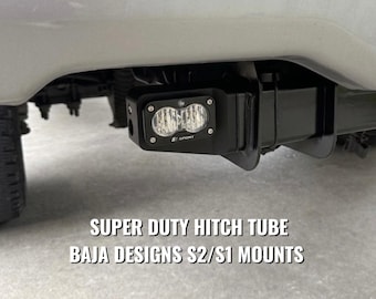 May include: A black metal hitch tube mount installed on a vehicle, featuring a rectangular Baja Designs S2 Sport LED light with a clear lens. The text SUPER DUTY HITCH TUBE BAJA DESIGNS S2/S1 MOUNTS is displayed in white letters below the light fixture.