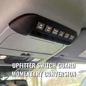 May include: A black overhead console upfitter switch panel installed in a vehicle ceiling. The panel features six labeled buttons: AIR HORN, HIGH IDLE, TRAILER CAMERA, AIR COMP, DITCH LIGHTS, and REVERSE LIGHTS, with a momentary conversion guard.