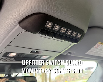 May include: A black overhead console upfitter switch panel installed in a vehicle ceiling. The panel features six labeled buttons: AIR HORN, HIGH IDLE, TRAILER CAMERA, AIR COMP, DITCH LIGHTS, and REVERSE LIGHTS, with a momentary conversion guard.