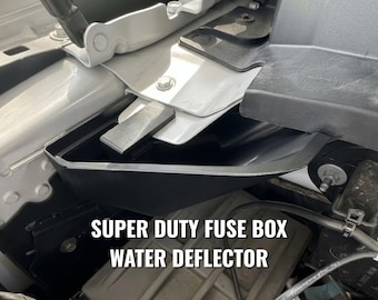 May include: A black plastic Super Duty fuse box water deflector installed in a vehicle engine bay. The component is positioned near the white metal frame and black plastic housing, featuring a molded shape designed to divert water away from the fuse box.