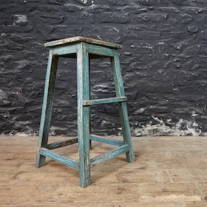 May include: A tall, four-legged wooden stool featuring a weathered, distressed teal blue paint finish. The stool has a square seat and horizontal support bars connecting the legs on all sides, showcasing a worn texture and visible wood grain.