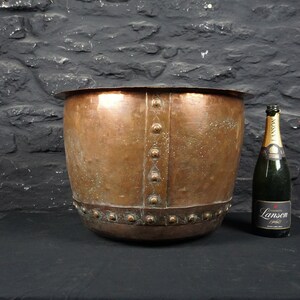 May include: A large copper planter with a hammered finish and decorative rivets around the top rim. The planter is sitting on a black surface.