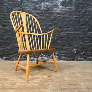 May include: A light-coloured wooden armchair with a curved back and armrests. The chair has a spindle back design and is set against a dark, textured wall. The chair's legs are angled for stability.