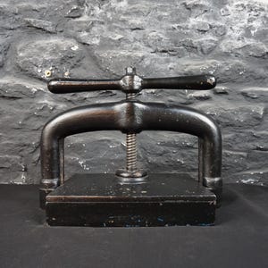 May include: A vintage black cast iron book press featuring a heavy rectangular base, two sturdy vertical supports, and a central threaded screw mechanism topped with a horizontal handle for applying pressure to bound books or paper documents.