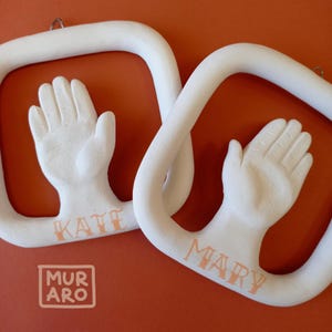 Puede incluir: Dos adornos de cerámica blanca con forma de huella de mano con los nombres "Katie" y "Mary" inscritos en letras naranjas. Los adornos están colgados de una cuerda blanca y tienen un fondo blanco.