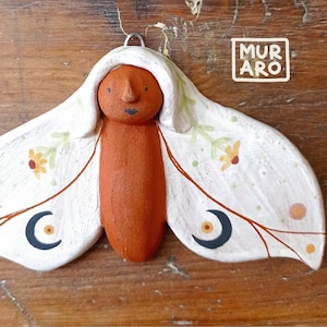 Könnte beinhalten: Eine Wanddekoration aus Terrakotta-Keramik in Form eines Nachtfalters mit weißen Flügeln. Die Flügel sind mit gemalten Blumen, Mondsicheln und Ästen verziert. Der Nachtfalter hat ein braunes Gesicht mit schwarzen Augen und einem schwarzen Mund.