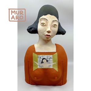 May include: A ceramic bust sculpture of a woman with black hair and yellow eyes. The woman wears an orange top with a painted window scene. The sculpture has a matte finish and a unique artistic style. The logo "MURARO" is in the upper left corner.