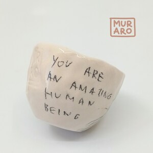 Puede incluir: Un pequeño cuenco de cerámica blanca con las palabras "You are an amazing human being" escritas en marcador negro.
