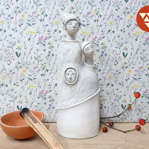 May include: White ceramic sculpture of two figures with stylised faces. The figures are standing on a wooden surface with a small bowl and a wooden stick.