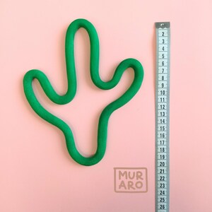 Könnte beinhalten: Eine grüne, kaktusförmige Tonskulptur auf rosa Hintergrund. Rechts neben der Skulptur befindet sich ein Lineal mit den Zahlen 2 bis 26.