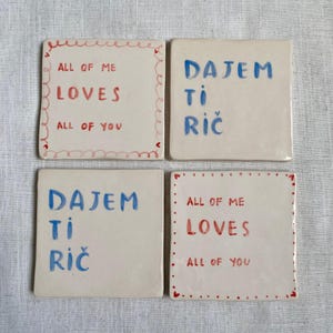 Può includere: Quattro sottobicchieri quadrati in ceramica con testo scritto a mano. Due sottobicchieri recitano "ALL OF ME LOVES ALL OF YOU" in rosso e nero. Gli altri due recitano "DAJEM TI RIČ" in blu. Ogni sottobicchiere ha un bordo decorativo.