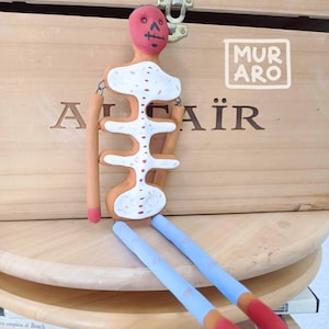 Puede incluir: Una muñeca de esqueleto de arcilla hecha a mano con una cara roja, huesos pintados de blanco y rojo y pantalones azules. La muñeca está sentada sobre una superficie de madera con el texto "MURAFAIR" en la superficie.