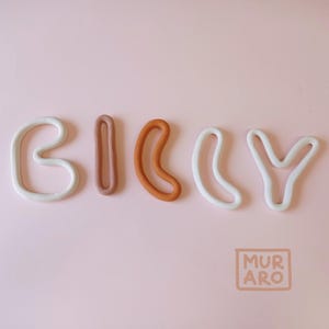 Puede incluir: El nombre "BILLY" escrito en letras de arcilla blanca, marrón y naranja. Las letras son estilizadas y tienen una forma redondeada y orgánica. Las letras están dispuestas en línea sobre un fondo rosa. El texto "MURARO" está en un cuadrado en la esquina inferior derecha de la imagen.
