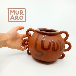 May include: A terracotta pot with a brown glaze and multiple handles. The pot has a wavy design in brown glaze.