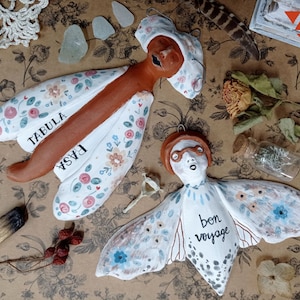 Puede incluir: Dos decoraciones de pared de cerámica hechas a mano con forma de mariposas. La primera mariposa es marrón con detalles florales blancos y rosas y el texto "TABULA RASA". La segunda mariposa es blanca con detalles azules y marrones y el texto "bon voyage".