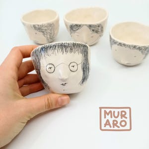 Può includere: Set di quattro tazze in ceramica bianca con volti disegnati a mano. Ogni tazza presenta un'espressione diversa, tra cui un viso con gli occhiali. Le tazze sono tutte leggermente diverse per dimensioni e forma. Le tazze sono firmate "MURARO" nell'angolo in basso a destra.