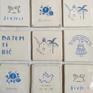 Op de afbeelding: Negen vierkante keramische tegels met blauwe handgetekende ontwerpen en tekst. Ontwerpen omvatten bloempatronen, een kerk met palmbomen, een duif, champagneglazen en een bruidstaart. Tekst bevat namen en zinnen zoals "better together".